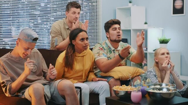 Seven friends sitting on a sofa watching a sports match at home during daytime, showing surprise and concentration while sharing snacks in a casual living room setting.