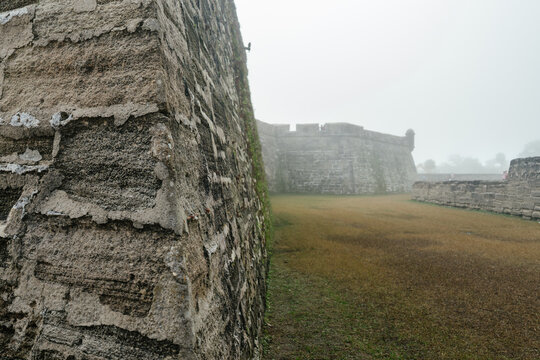 A close stone wall overlooks the grassy courtyard of Castillo de San Marcos. Thick fog surrounds the historic coquina bastions and creates a mysterious atmosphere.