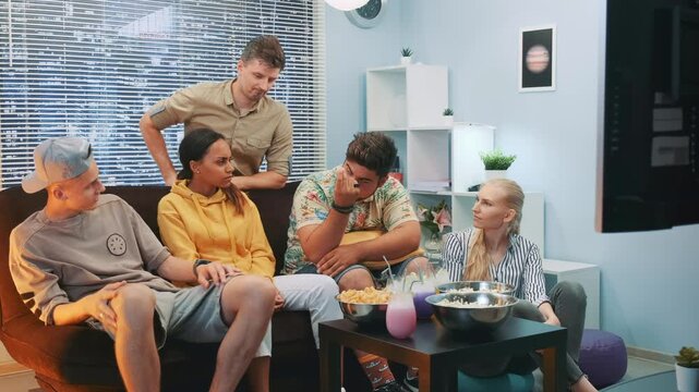 Six adults casually gathered around a coffee table with snacks and drinks in a bright, modern indoor living room during daytime, enjoying a relaxed social moment.