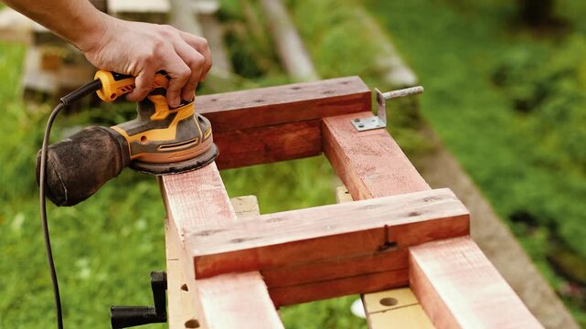 4K slow motion 60 fps footage of a craftsman hand using an electric orbital sander tool to smooth a brown wooden construction frame outdoors on a sunny day.