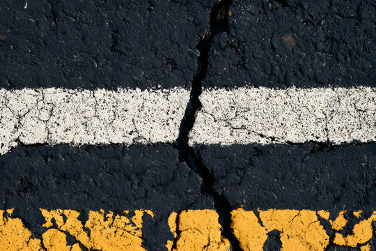 A close-up view reveals the texture of the old asphalt road. White and yellow crack lines are visible on the dark, rough street surface. The weathered bitumen displays damaged markings, creating a gru