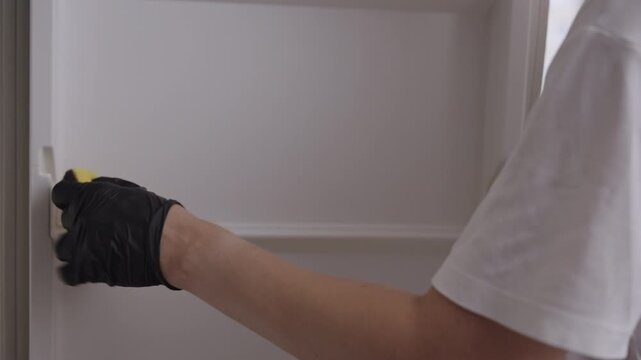 Homemaker in black gloves thoroughly cleaning and disinfecting interior of empty refrigerator door with steam generator and sponge for fresh and hygienic kitchen. Concept of household chores.