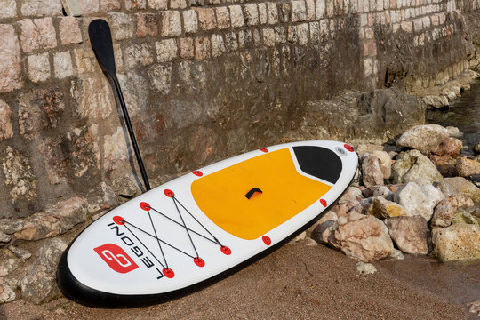 July 2, 2026: Budva, Montenegro, Close view of stand up paddleboard on pebbled shoreline next to weathered concrete seawall, paddle propped against wall with seaweed and stones around it in bright sun