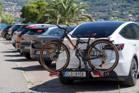 July 4, 2026, Kotor, Montenegro, White hatchback fills clear foreground while mountain bike sits on rear rack, with parked cars receding toward coastal town, palm trees, hillside buildings, and soft s