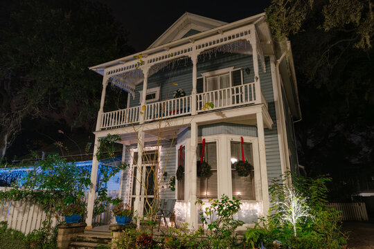 A blue Victorian house with white balconies and columns stands illuminated at night in St. Augustine, Florida. Christmas lights and wreaths decorate the windows, porch, and tropical garden.