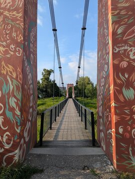 Ornate carved wooden gateway framing narrow suspension footbridge stretching over green riverside park under clear blue sky