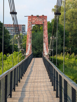 Weathered pedestrian suspension bridge with wooden planks and rusted metal cables leading toward green riverside park under soft summer daylight