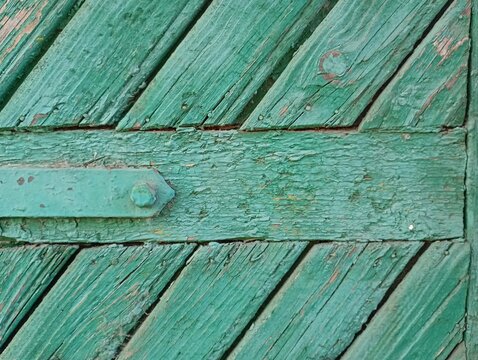 A close up texture of an old weathered wooden door painted in green, featuring diagonal planks, a horizontal beam with a rusty metal hinge, and heavily peeling paint details.