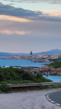 Vertical view of the Alghero shoreline at sunset, with the Mediterranean Sea in the foreground and the city of Alghero visible in the distance.