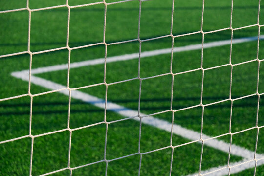 White soccer goal net viewed closely against vivid green artificial turf with intersecting field markings and geometric mesh pattern in outdoor sports field setting