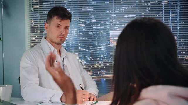 A male family consultant in a white coat listens and takes notes while a woman speaks expressively in a nighttime office with city skyscrapers visible outside.