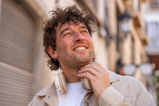Young man with curly hair wearing headphones around neck, smiling confidently outdoors, enjoying urban street life