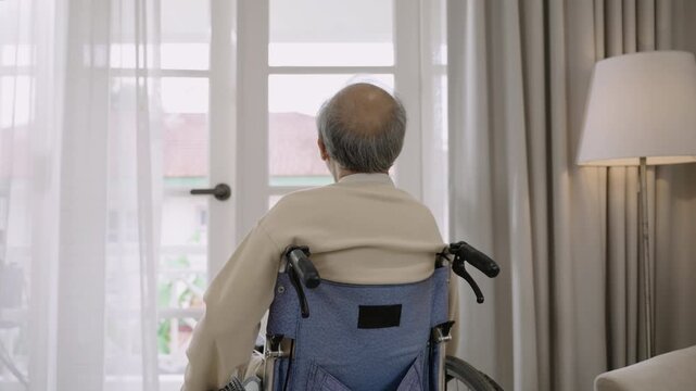 A senior Asian man is sitting alone in wheelchair and looking outside window due to depression and loneliness in his room at home. Sad elderly old male patient is lonely. 