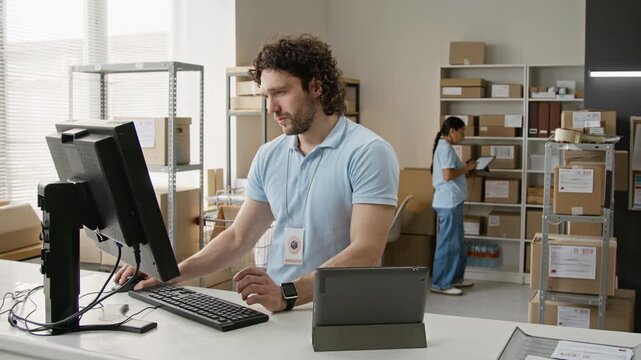 Caucasian young man managing orders on computer and tablet at fulfillment warehouse counter. Ideal footage for e-commerce logistics, supply chain software, and inventory management campaigns