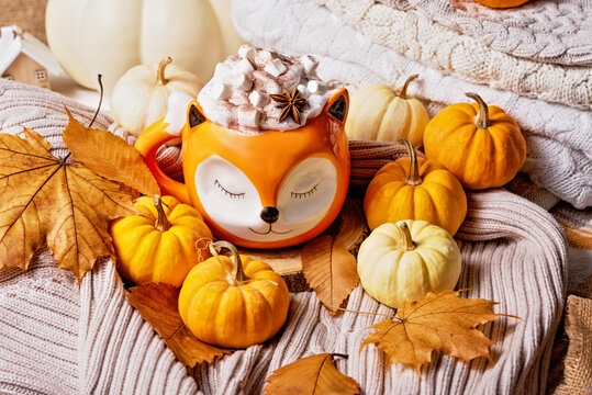 Creamy pumpkin spice latte topped with marshmallows and spices in Cute orange fox mug, surrounded by pumpkins and colorful autumn leaves.