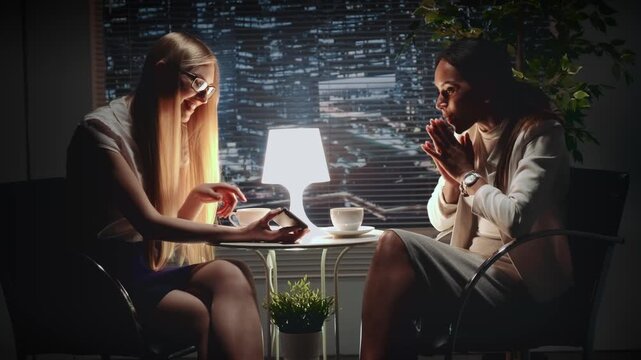 Two businesswomen sit at a small table in a cafe at nighttime, one focused on a smartphone while the other listens, with a city skyline visible through a large window.