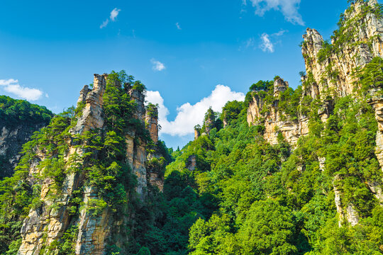 Vertical limestone karst cliffs rise above a dense green forest, with rugged rock pillars framing a shadowed valley beneath blue sky and scattered clouds.