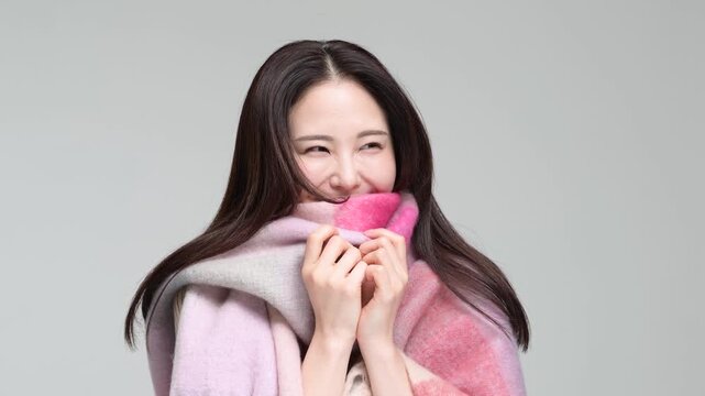 グレー背景の室内でマフラーに包まりながら笑顔を見せる30代の日本人女性
