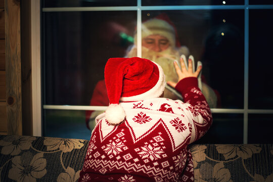 Cute boy in a Santa hat looking out the window. An authentic Santa Claus stands outside.