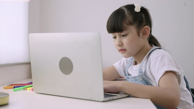 Cute Asian girl student sitting at desk using a laptop computer for studying online learning via video calling with remote teacher in a home school. Homeschooling education and new normal concept. 