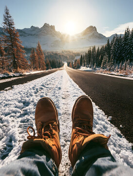 POV shot of man's brown leather boots walking on snowy road. Traveler explores winter mountain landscape under bright sun. Man stands through frost on cold highway with pine trees and snowy peaks in t