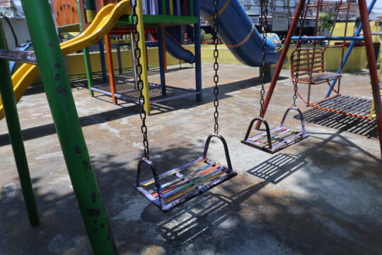 Colorful metal swings and slides in a public playground in Java, Indonesia. The empty playground equipment is illuminated by sunlight, casting distinct shadows on the concrete ground.