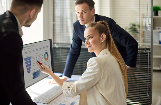 Woman analyst with colleagues reviewing data. She points to charts on a computer screen as colleagues plan strategy, review KPIs, and prepare a report. Concept is team analytics in office.