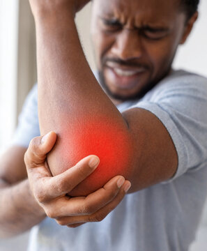 Adult Black man holds his painful elbow with a red highlighted area, showing joint pain or injury in an indoor setting.
