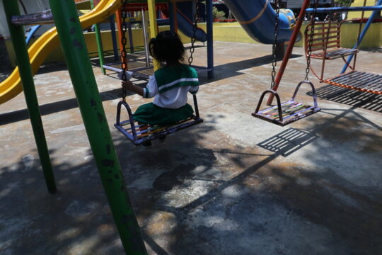 A kindergarten child wearing a school uniform plays alone on a swing at a public playground after school. Colorful playground equipment, sunlight, trees, and shadows.