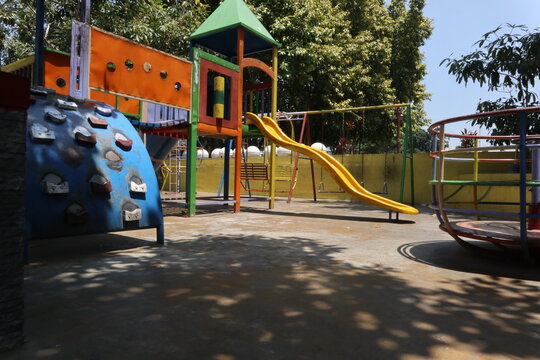 Colorful outdoor playground in a public park in Java, Indonesia, featuring a slide, swings, merry-go-round, climbing structure, and other recreational equipment under trees on a sunny day.