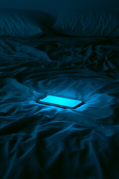 Minimalist glowing blue smartphone on dark bed with moody low key lighting cold cyan