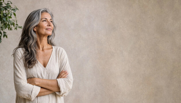 Mature woman with gray hair stands with crossed arms against a neutral wall, looking away with a calm, confident expression and copy space.