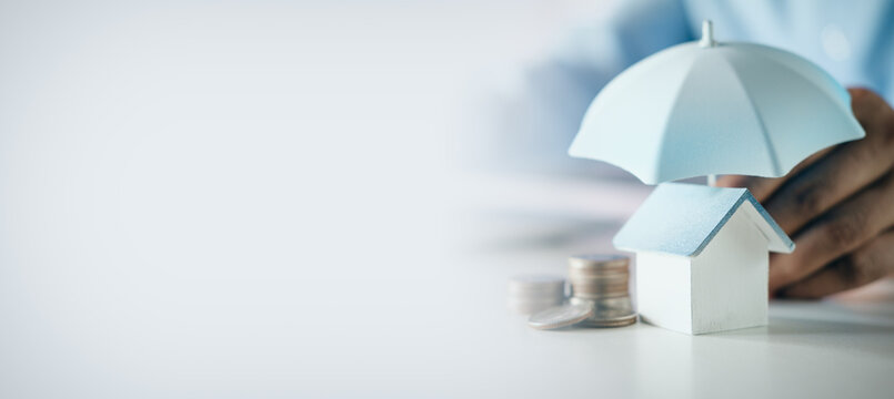 Hand holding a blue umbrella protecting a wooden house model and coin stacks on desk with copy space, concept of home insurance, property protection, security, and mortgage planning.
