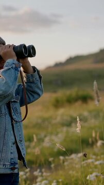 Vertical video: Scanning boy in beanie denim jacket using binoculars in meadow at sunset