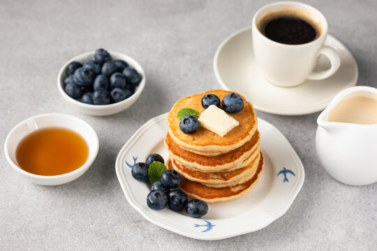 Golden fluffy pancakes topped with butter, blueberries and fresh mint on a white plate, accompanied by coffee and syrup on a light stone background.