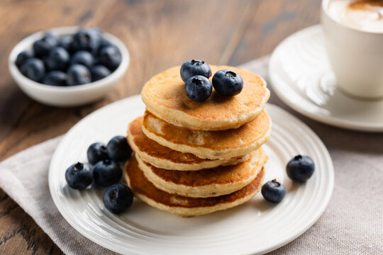 Stack of pancakes with blueberries on white plate served with cup of black coffee. Wooden table background