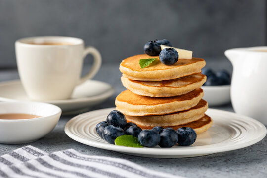 Golden fluffy pancakes topped with butter, blueberries and fresh mint on a white plate, accompanied by coffee and syrup on a light stone background.