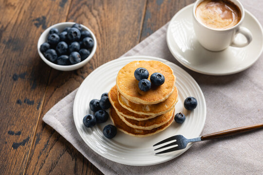 Stack of pancakes with blueberries on white plate served with cup of black coffee. Wooden table background