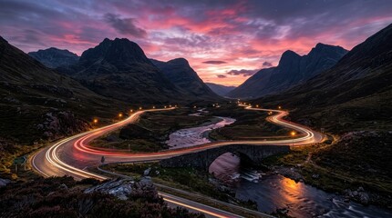 Obraz premium Dramatic Sunset over Glencoe Valley with Car Light Trails on a Winding Mountain Road and Stone Bridge in Scotland