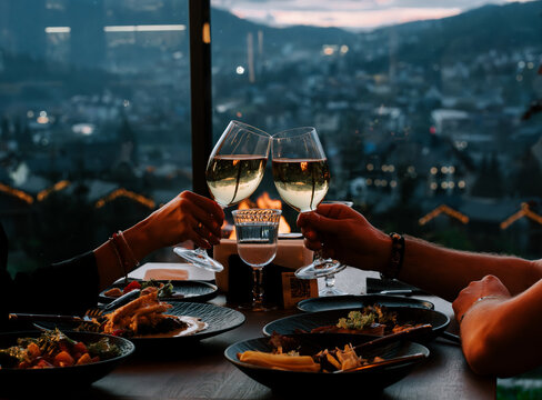 People toasting with white wine glasses during a romantic dinner overlooking a city at twilight