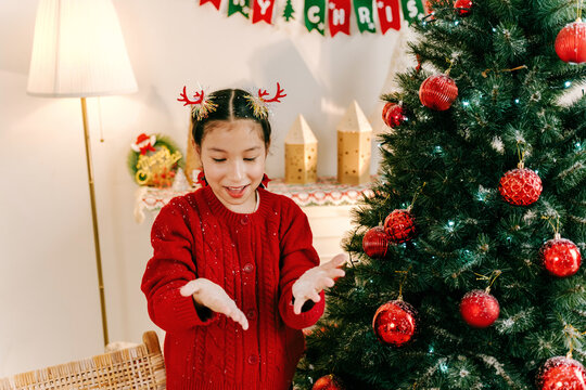 A child in a red sweater, wearing reindeer antlers, smiles and looks at her hands beside a decorated Christmas tree with red ornaments in a cozy, festive living room.