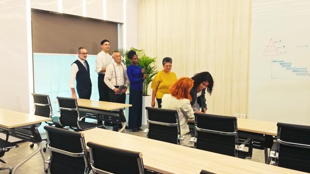 A diverse group in a modern training room lines up near desks and chairs to speak with a seated facilitator, preparing for a session with a whiteboard in an office setting.