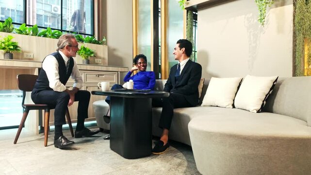 Three professionals converse around a coffee table in a modern office lounge with plants and city views, suggesting a collaborative business meeting