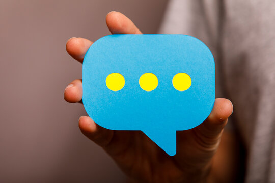 A person's hand holds a blue digital speech bubble with yellow dots, symbolizing online