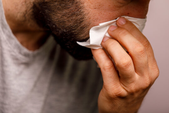 A close-up shot of a man's hand using a white tissue to clean his bearded cheek.