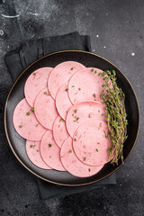 Sliced pink lunch meat (bologna/mortadella) with fresh thyme on a black plate. Delicious cold cut and charcuterie concept on a dark background. © Mironov