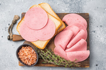 Deli meat and ingredients for a quick sandwich: pink processed ham, bread, herbs, and salt, on a dark wooden board and grey surface. © Mironov
