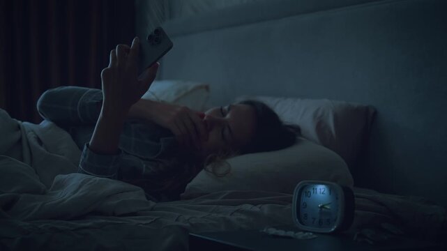woman suffers from insomnia lying in bed using smart phone before sleep, dark night room. bright light from affects eyes girl face. sad lady scrolling social media app phone screen holding mobile