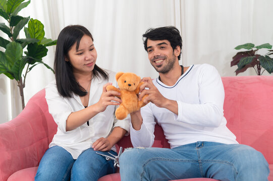 A joyful couple interacts with a teddy bear on a cozy pink couch, showcasing affection and playful interaction in a warm indoor setting.