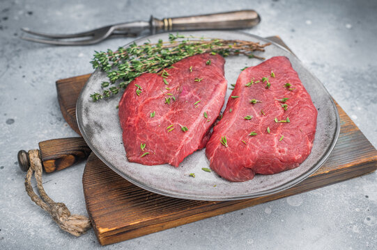 Raw Wagyu Top Blade chuck steaks with thyme on gray ceramic plate over light concrete background perfect for gourmet butcher and culinary stock photography.